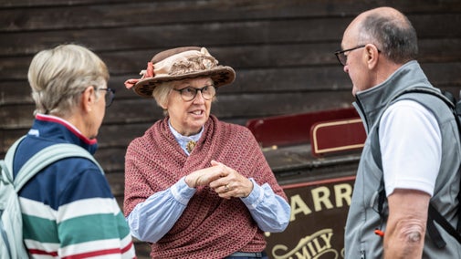 A woman in Victorian costume and hat speaks to two visitors to Blists Hill. She is wearing a rose coloured shawl and glasses.